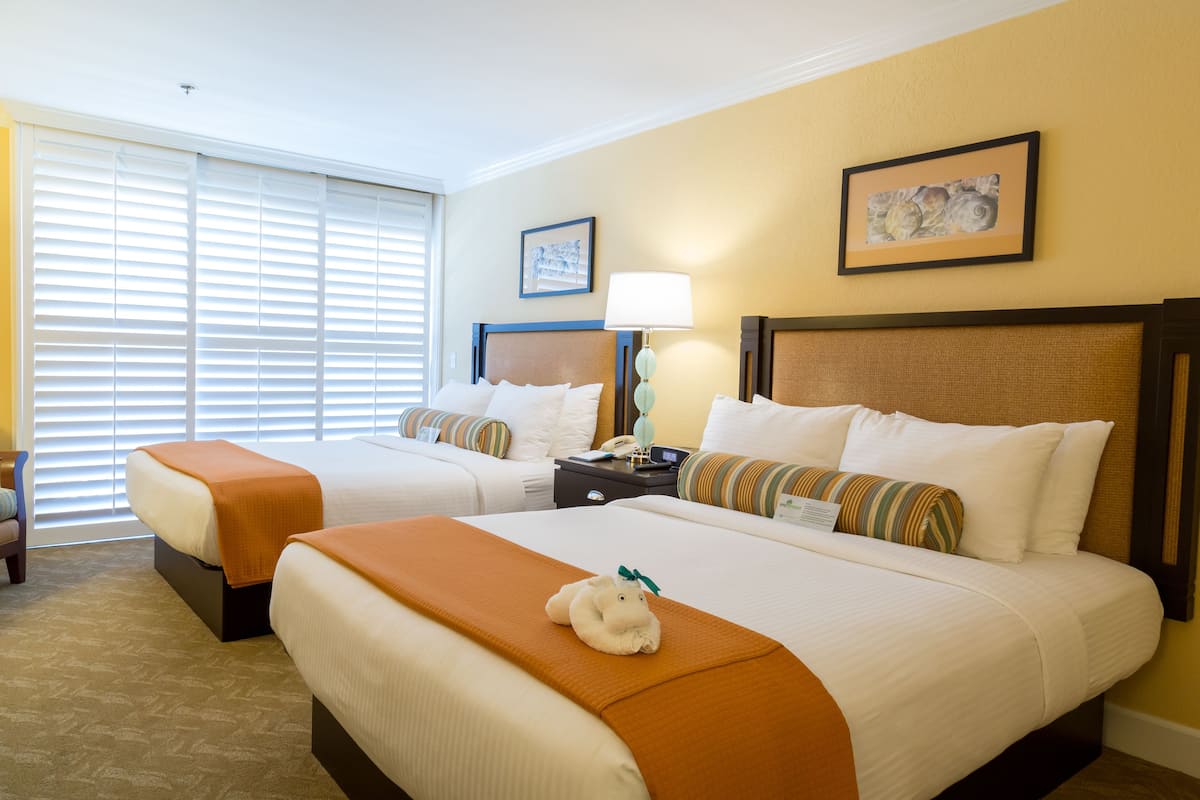 A hotel room with two beds, a nightstand with a lamp, a framed picture on the wall, and a window with blinds.