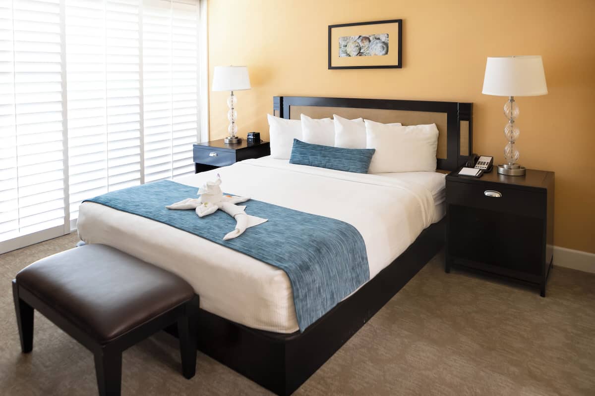 A hotel room with a large bed, two bedside tables, a bench, and a window with blinds.