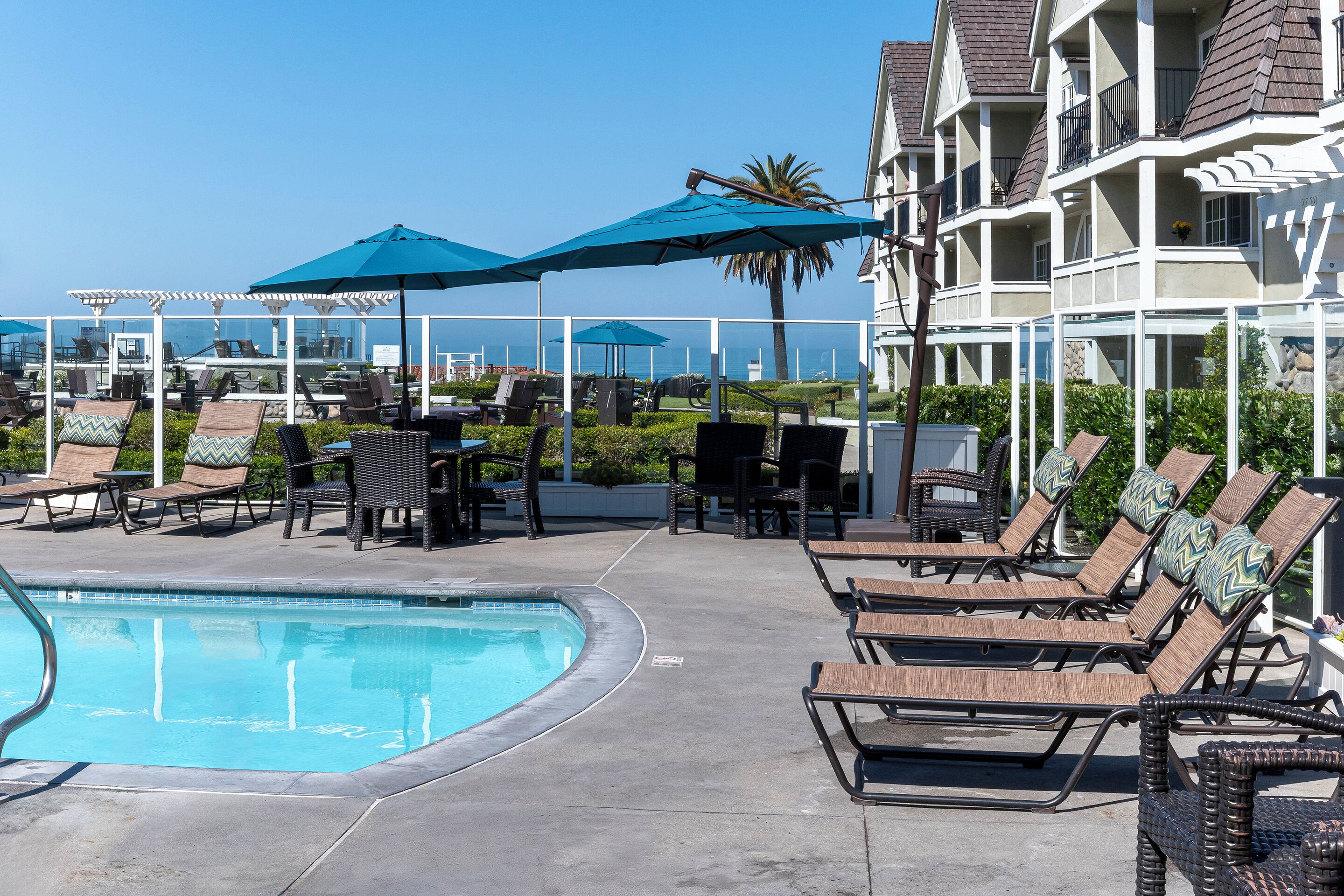 Un espace aménagé au bord de la piscine, avec des chaises longues et des parasols, une vue dégagée sur l’océan et un bâtiment doté de balcons.