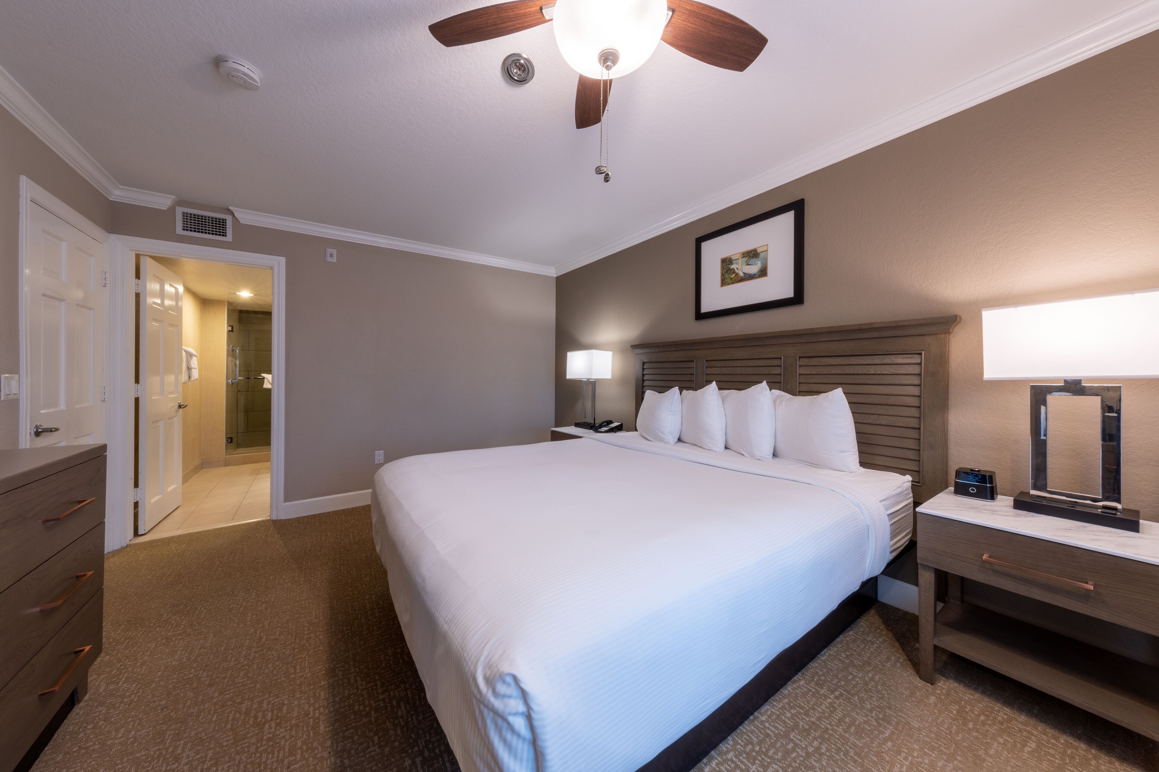 Een hotelkamer met een groot bed, een plafondventilator, een nachtkastje met een lamp, en een deur naar een badkamer.