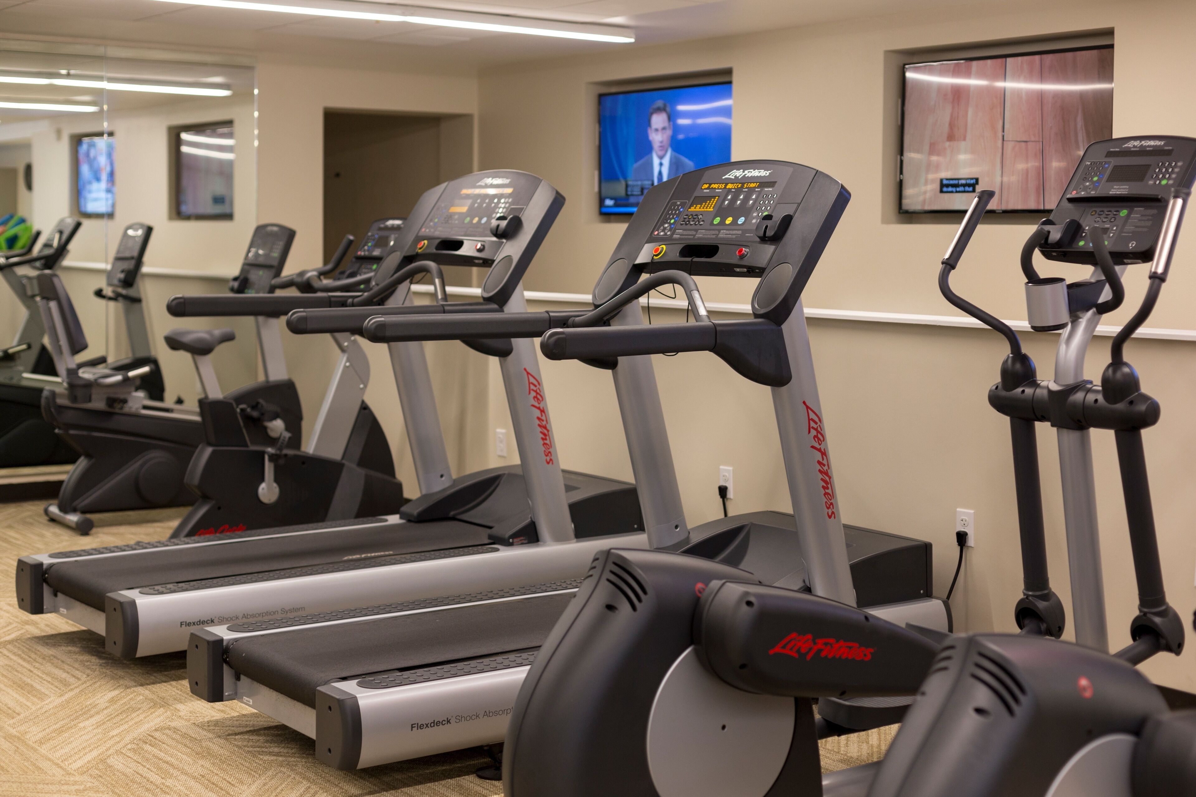 Varias cintas de correr en un gimnasio con un televisor colgado en la pared.