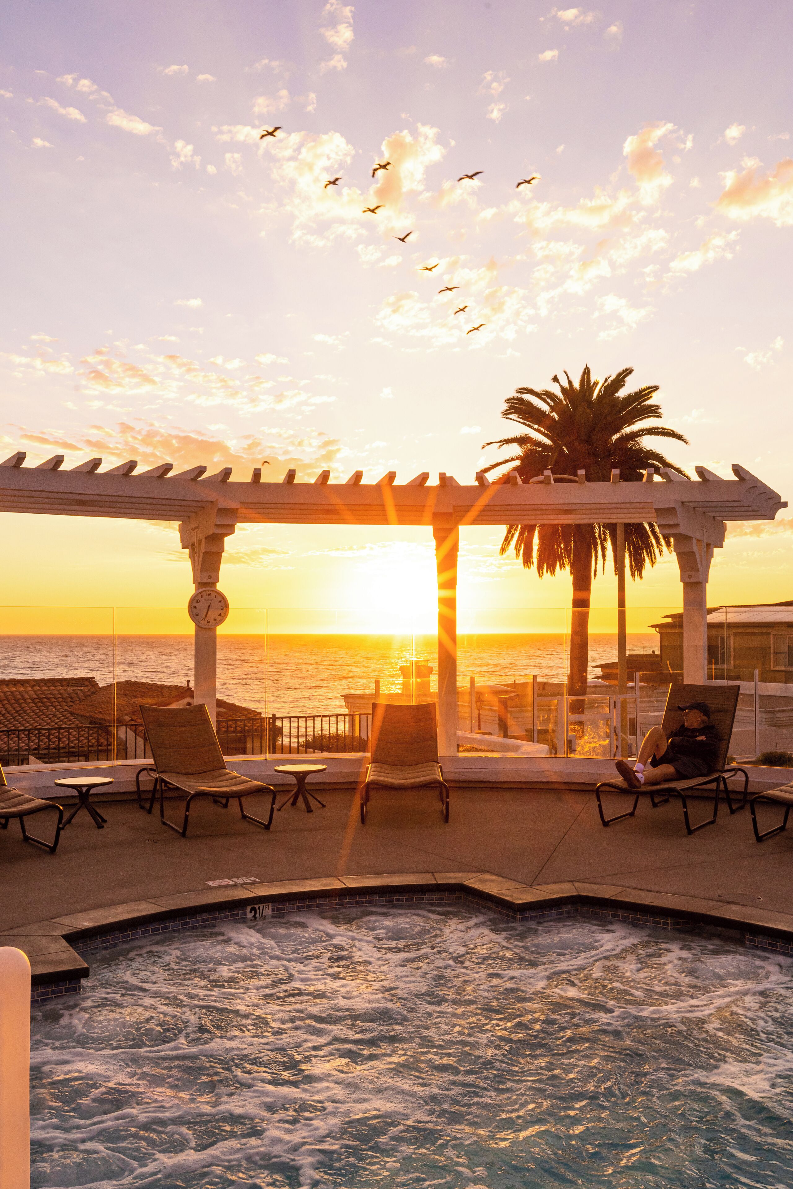 A poolside area with lounge chairs, a pergola, and a view of the sunset over the ocean.