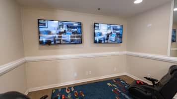 Ein Zimmer mit zwei großen, an der Wand befestigten Fernsehern, die Innenräume zeigen, einem schwarzen Massagesessel und einem gemusterten Teppich.