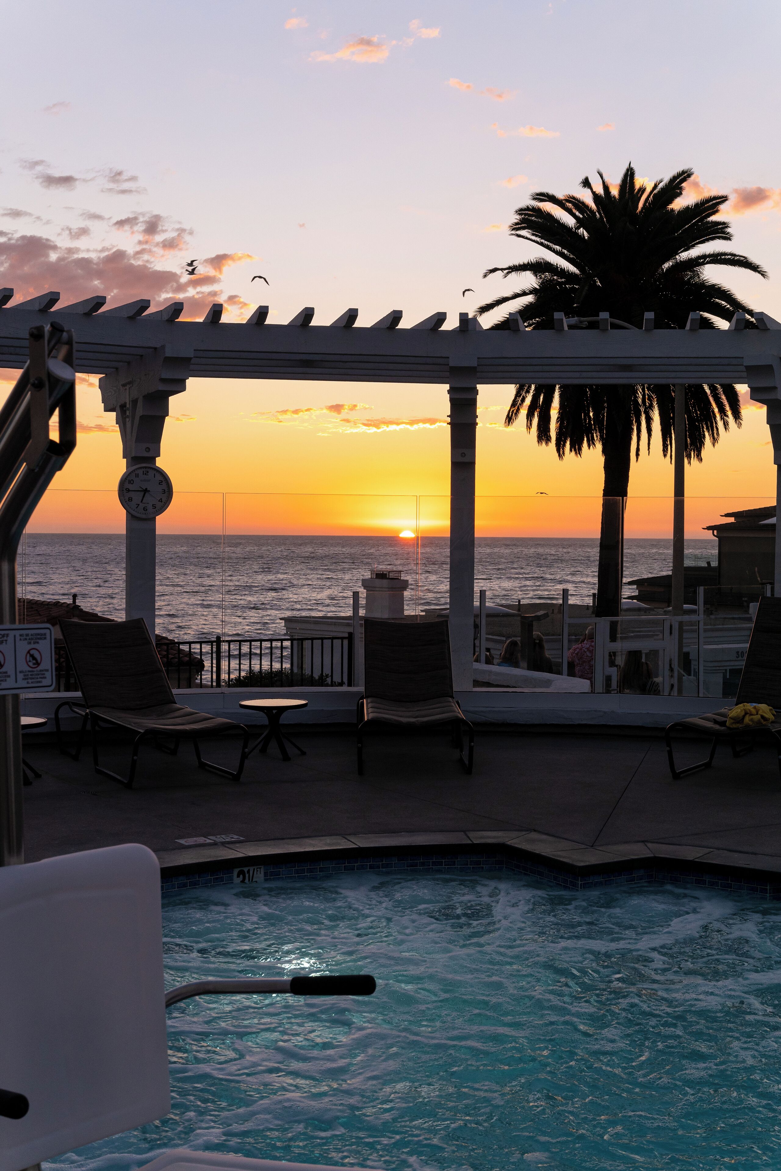 A pool area with lounge chairs and a view of the sunset over the ocean.
