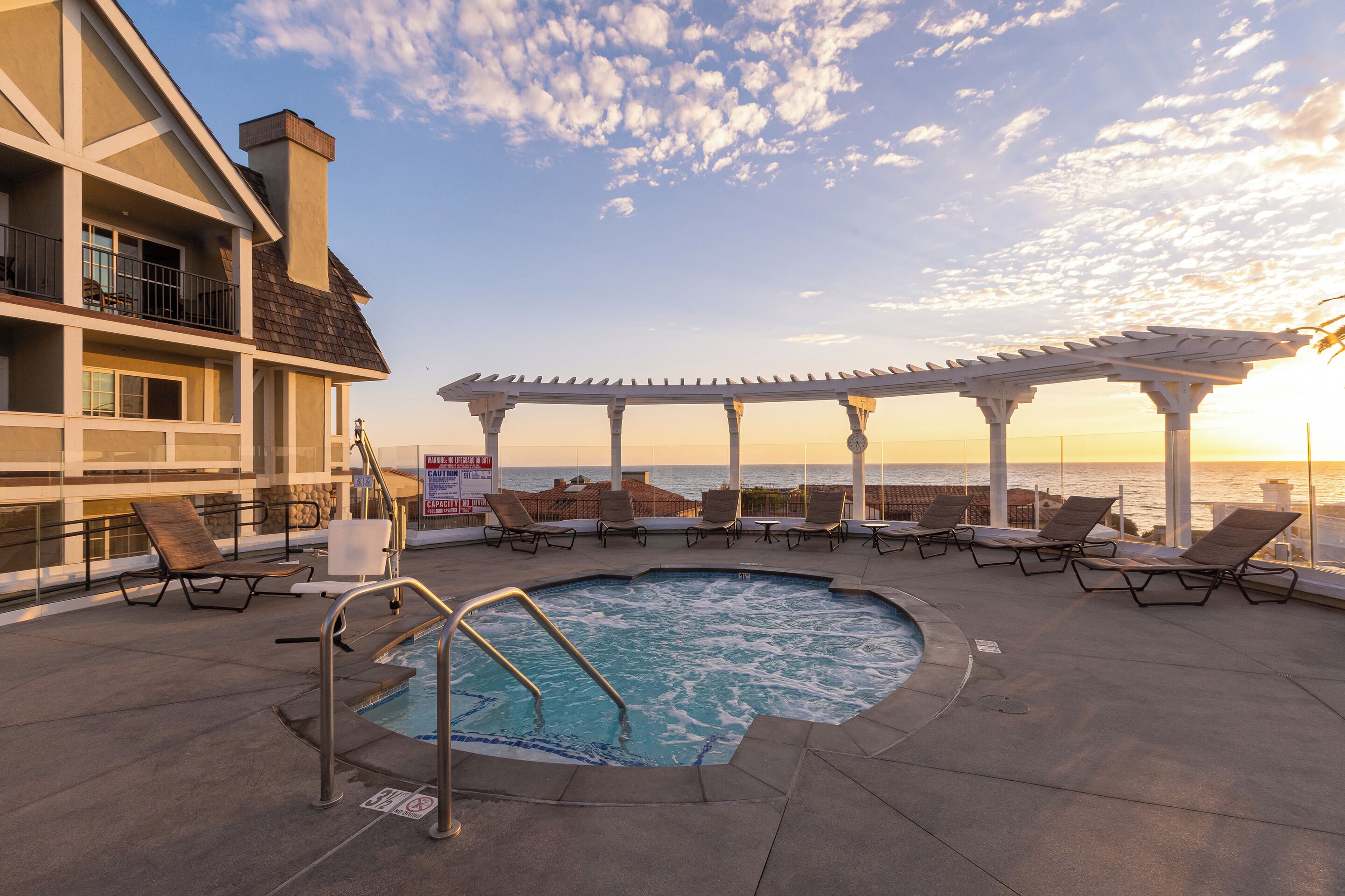 A rooftop pool area with lounge chairs, a pergola, and a view of the ocean at sunset.