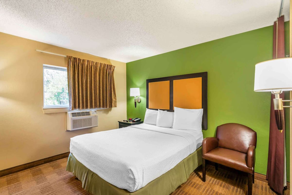 Ein Hotelzimmer mit einem großen Bett, einem Sessel, einem Fenster mit Vorhängen, einer Wand-Klimaanlage und einem Nachttisch mit Telefon und Lampe.