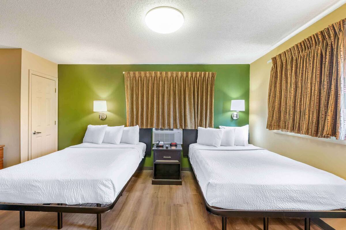 Ein Hotelzimmer mit zwei Betten, einem Flachbildfernseher, einem Schreibtisch mit Lampe und einem Fenster mit Vorhängen.
