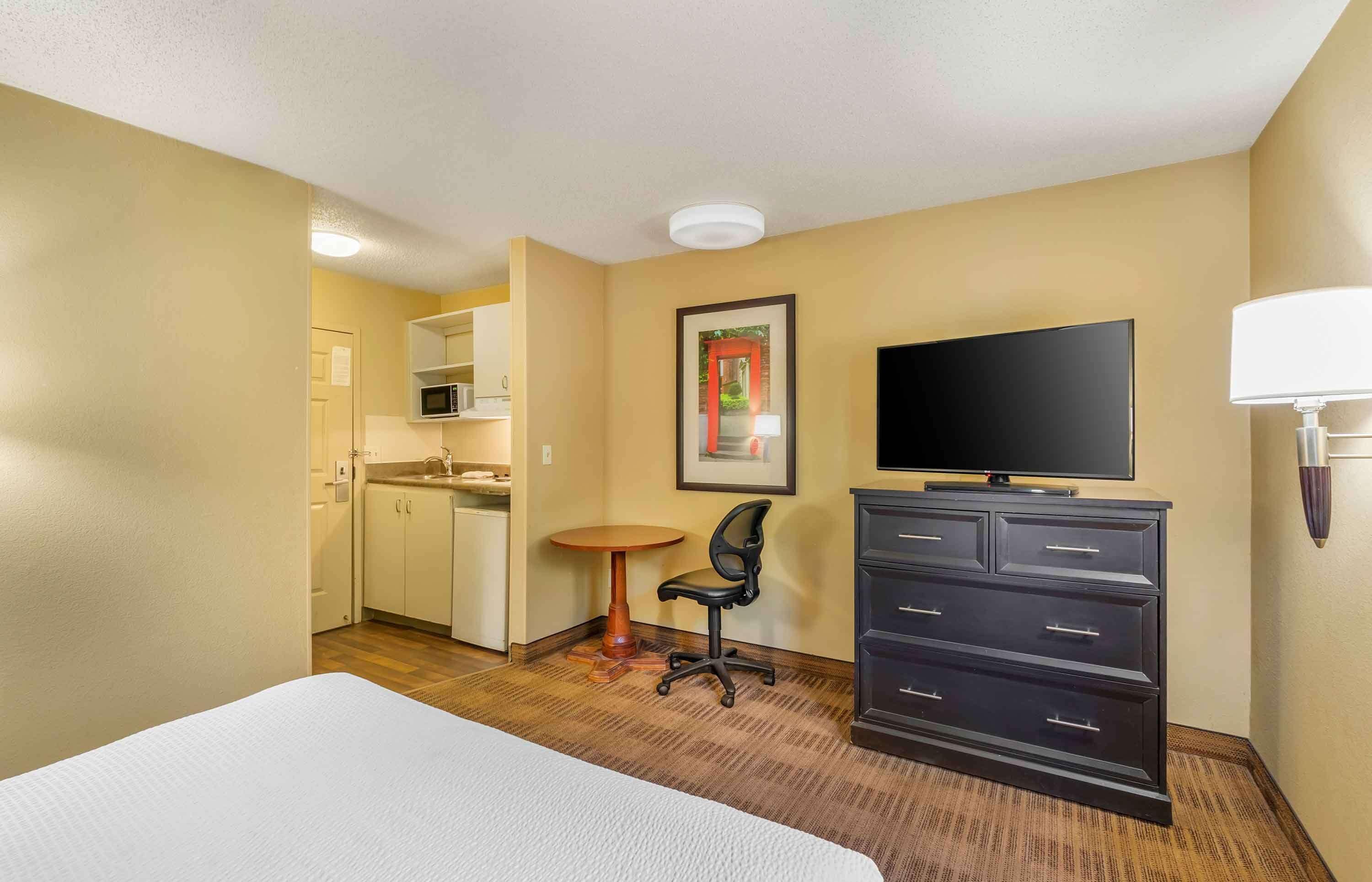 A hotel room with a bed, a desk with a chair, a TV, a dresser, and a microwave in the kitchenette.