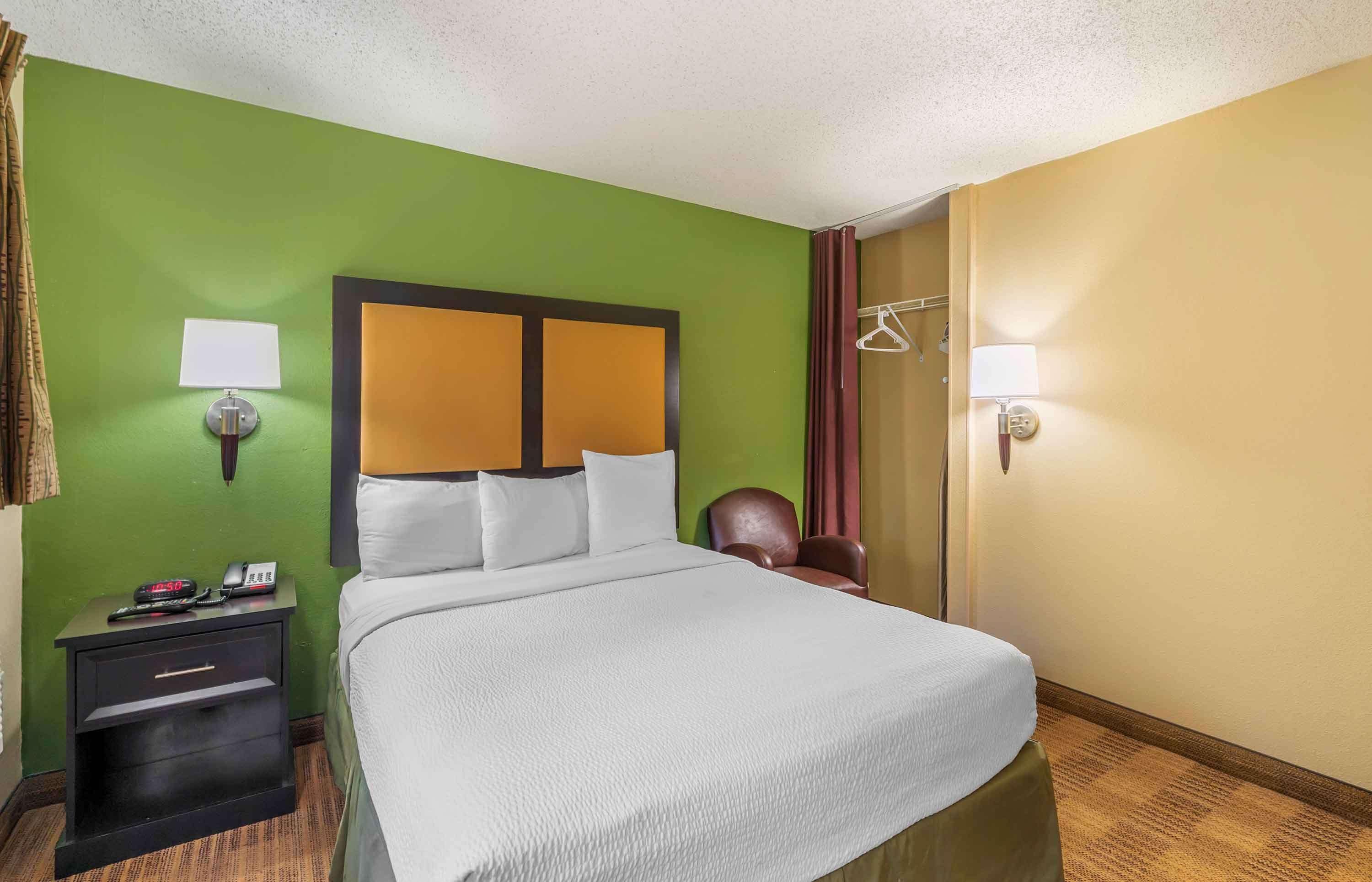 Quarto de hotel com cama, mesa de cabeceira, abajur e uma cadeira.