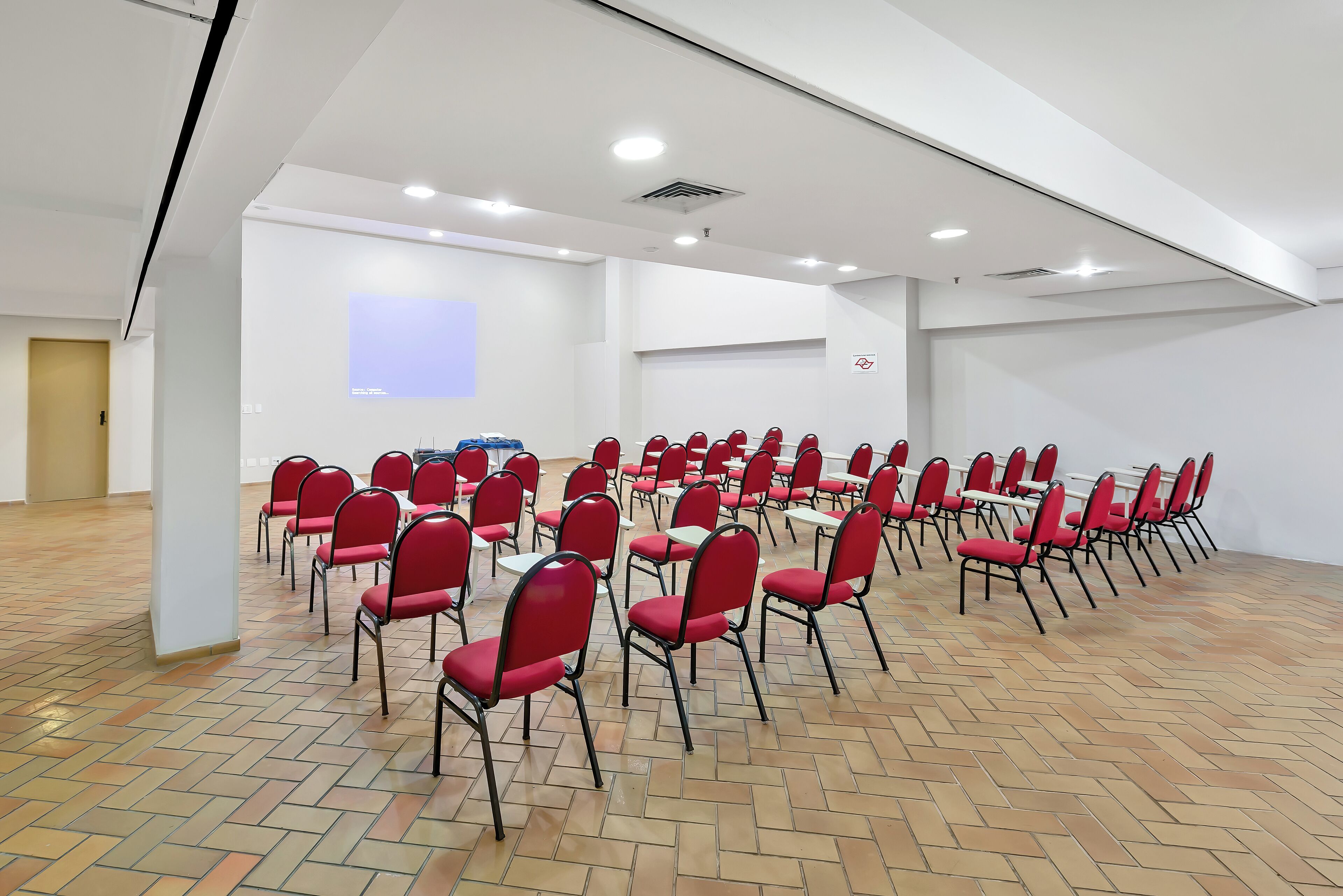 A conference room with rows of red chairs and tables, white walls, and a herringbone floor pattern.