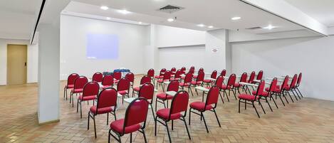 A conference room with rows of red chairs and tables, white walls, and a herringbone floor pattern.