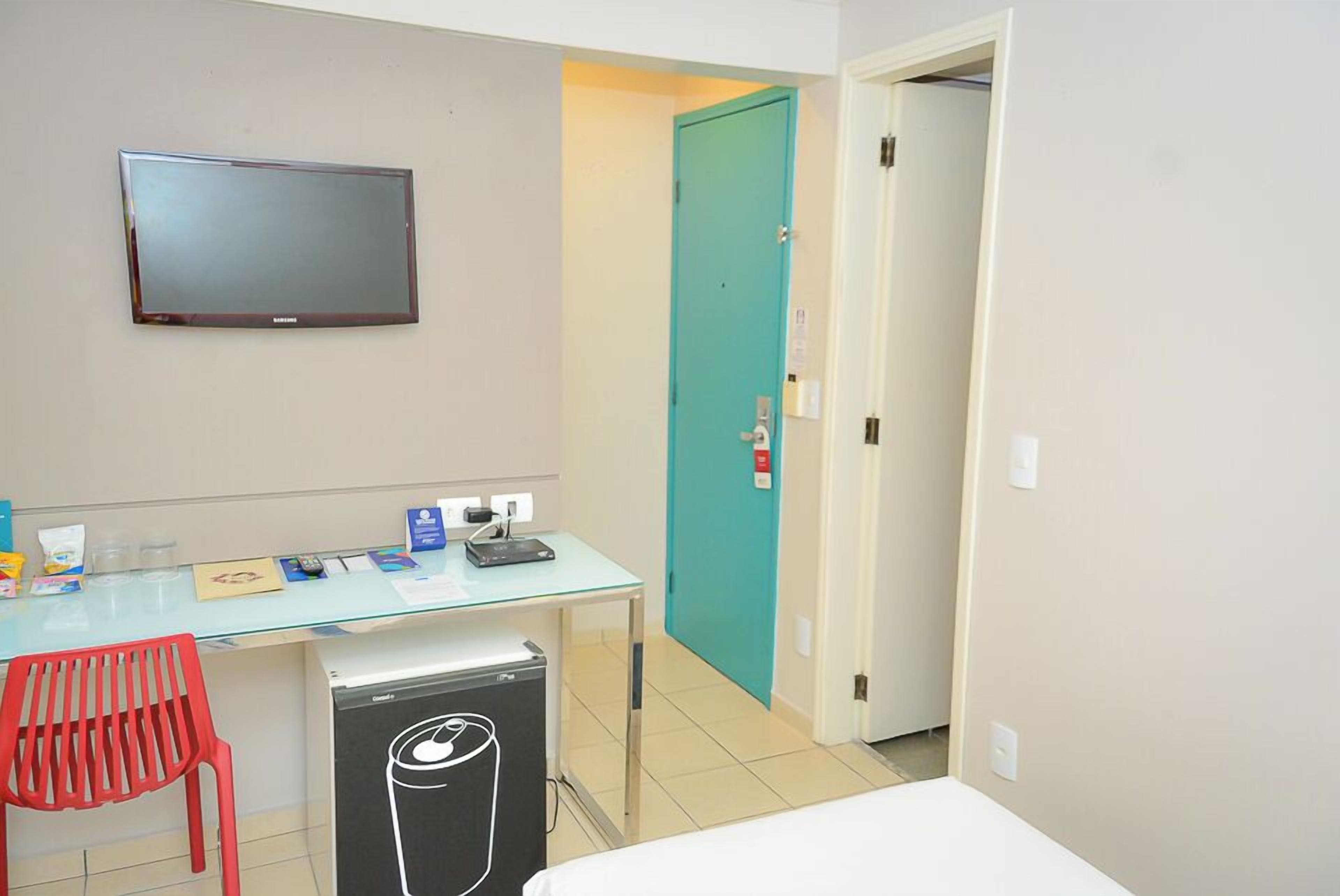 A hotel room with a flat-screen TV mounted on the wall, a desk with a computer and various items, a red chair, and a compact refrigerator.