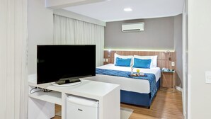 Habitación de hotel con cama, escritorio con televisor, mini refrigerador y mesita de noche con lámpara.