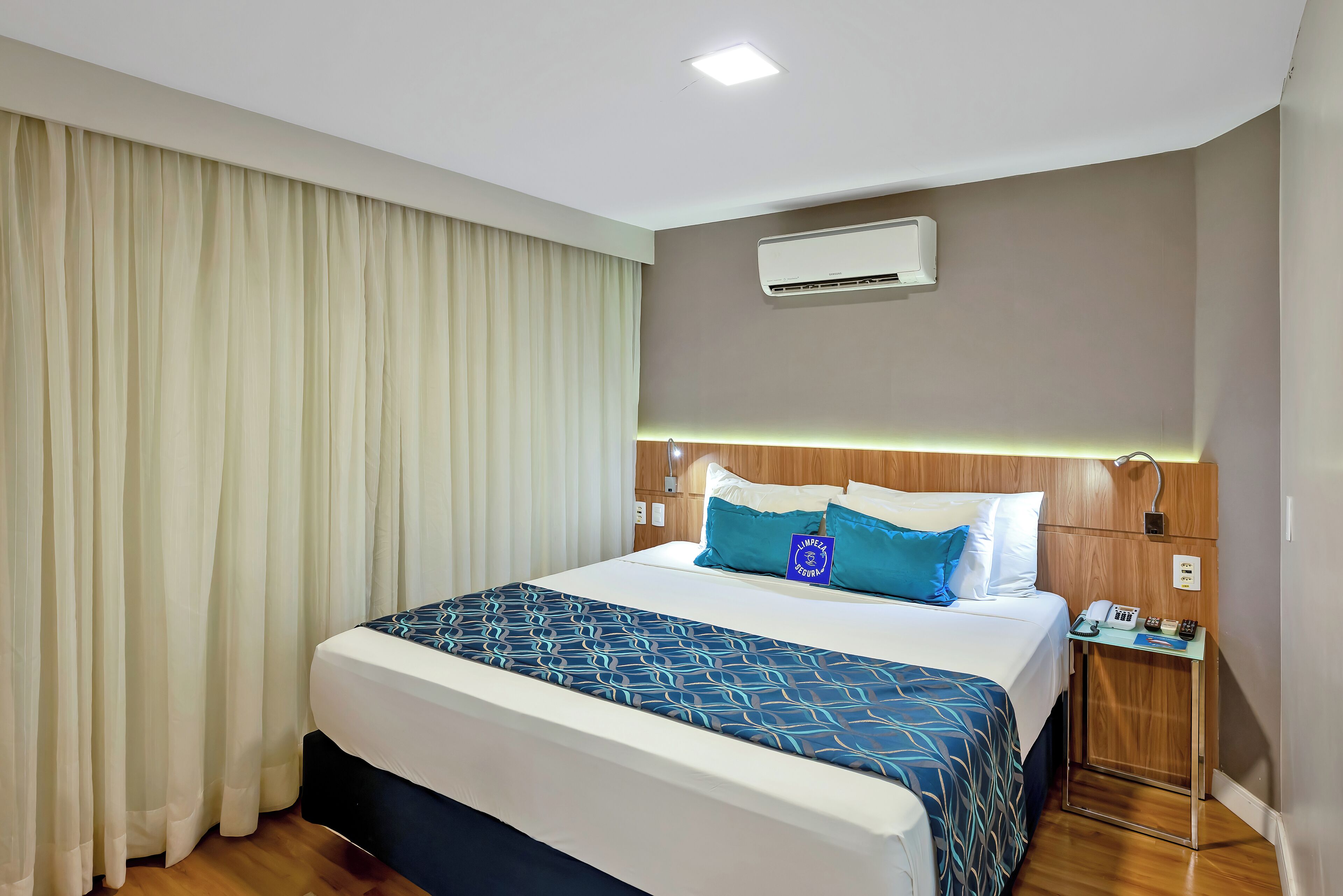 A hotel room with a large bed, a nightstand with a phone, and an air conditioning unit on the wall.