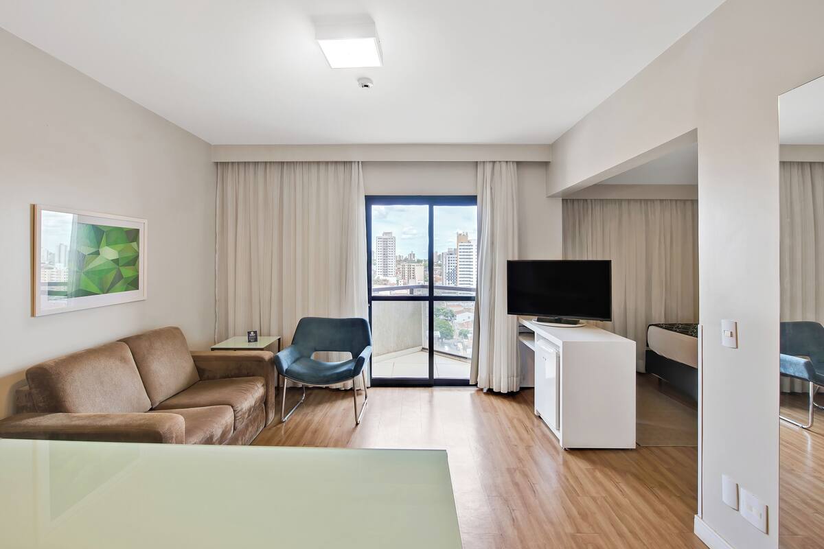 Habitación de hotel moderna con sofá, una silla, escritorio con televisor y ventana grande con vista a la ciudad.