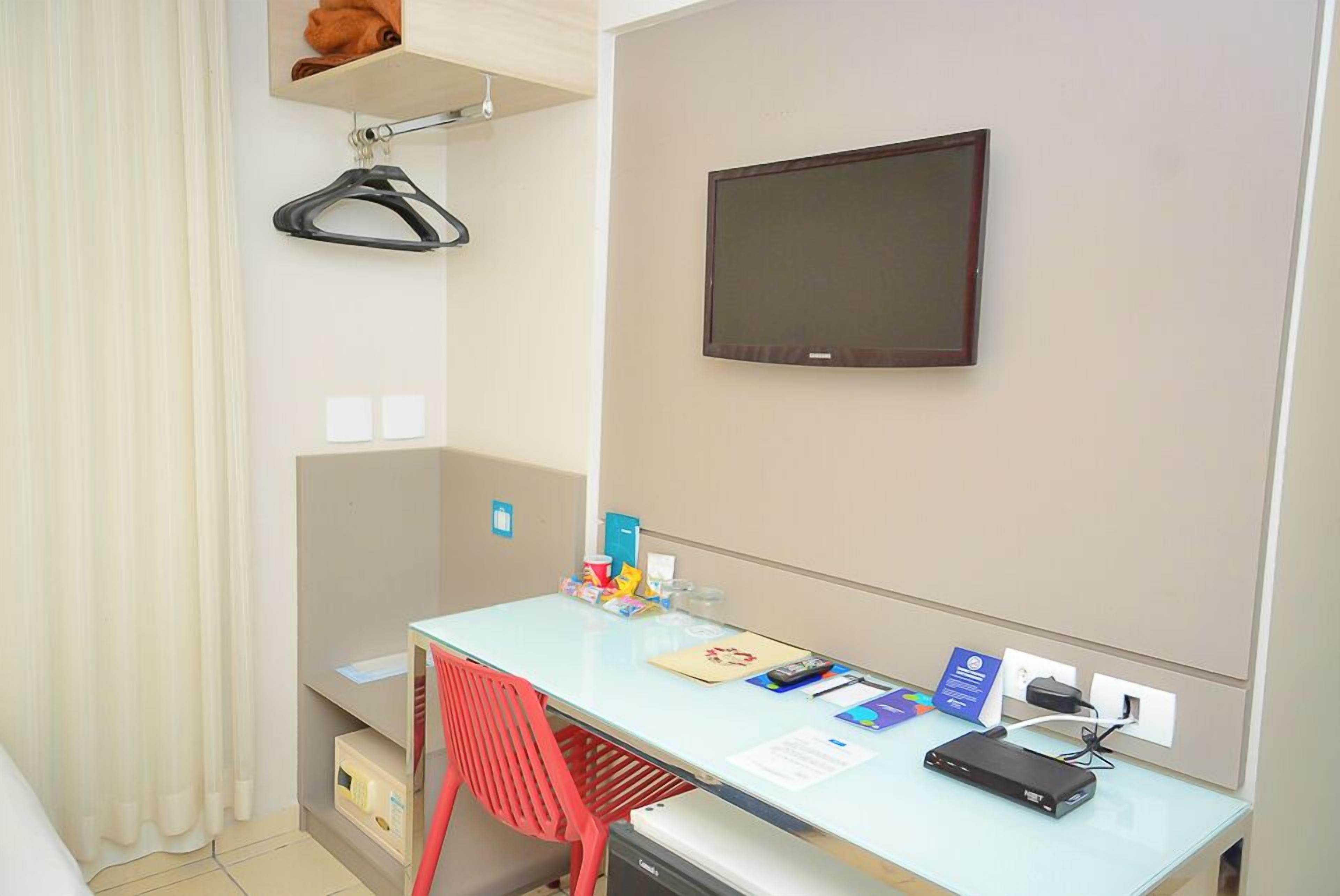 Ett hotellrum med ett skrivbord, en tv som är monterad på väggen, en röd stol och en klädställning med en galge.