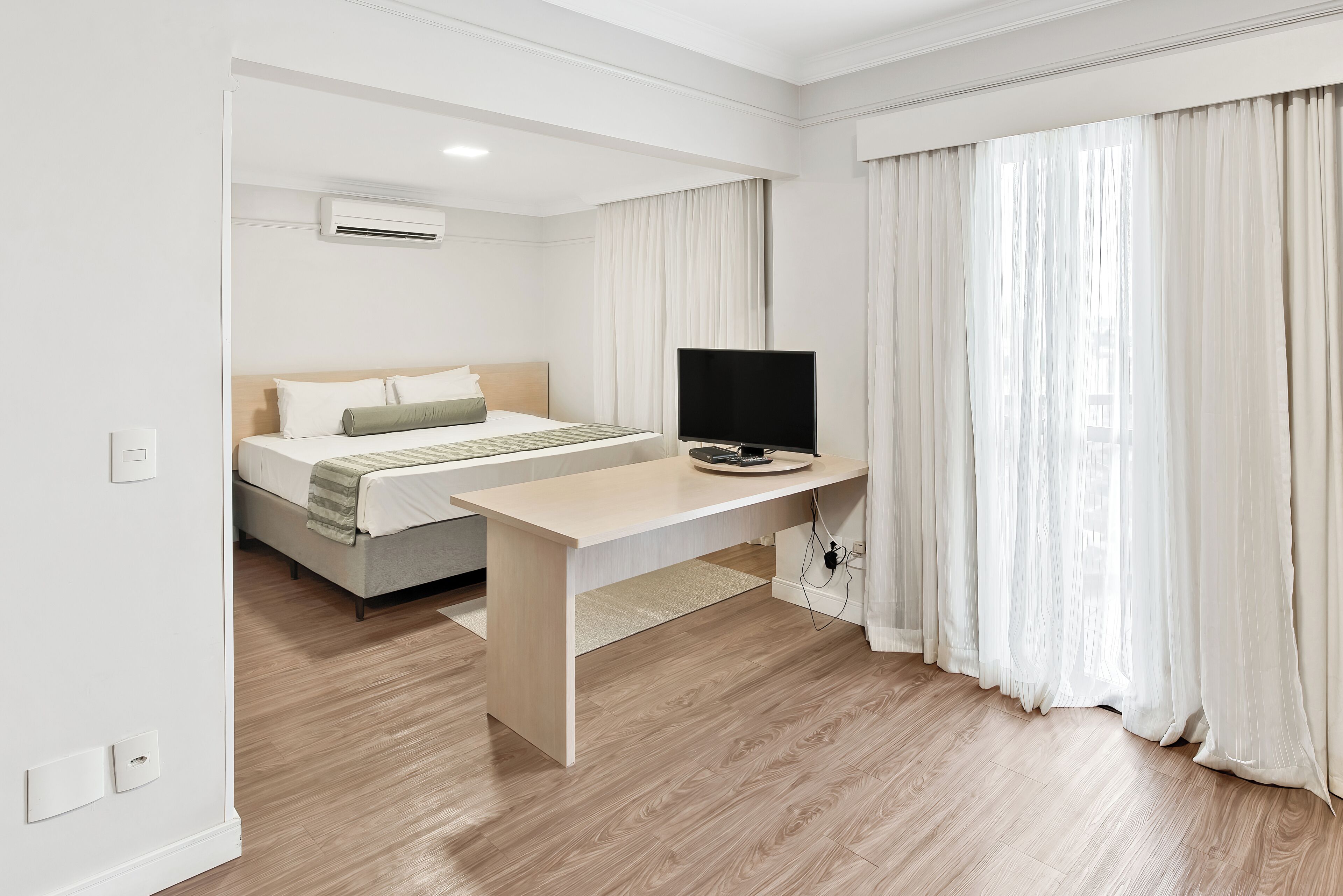 A hotel room with a bed, a desk with a TV, and a window with curtains.