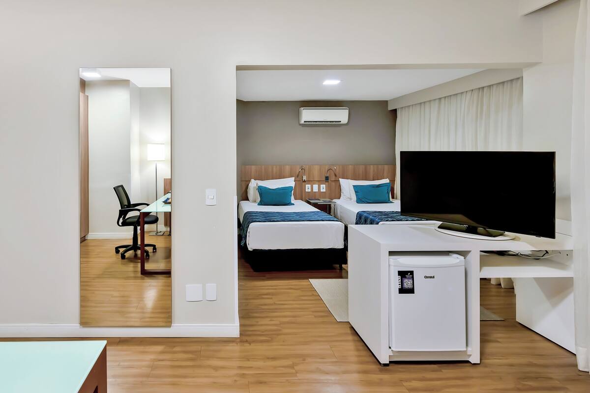 Habitación de hotel con dos camas, un escritorio con computadora, televisión y un refrigerador pequeño.