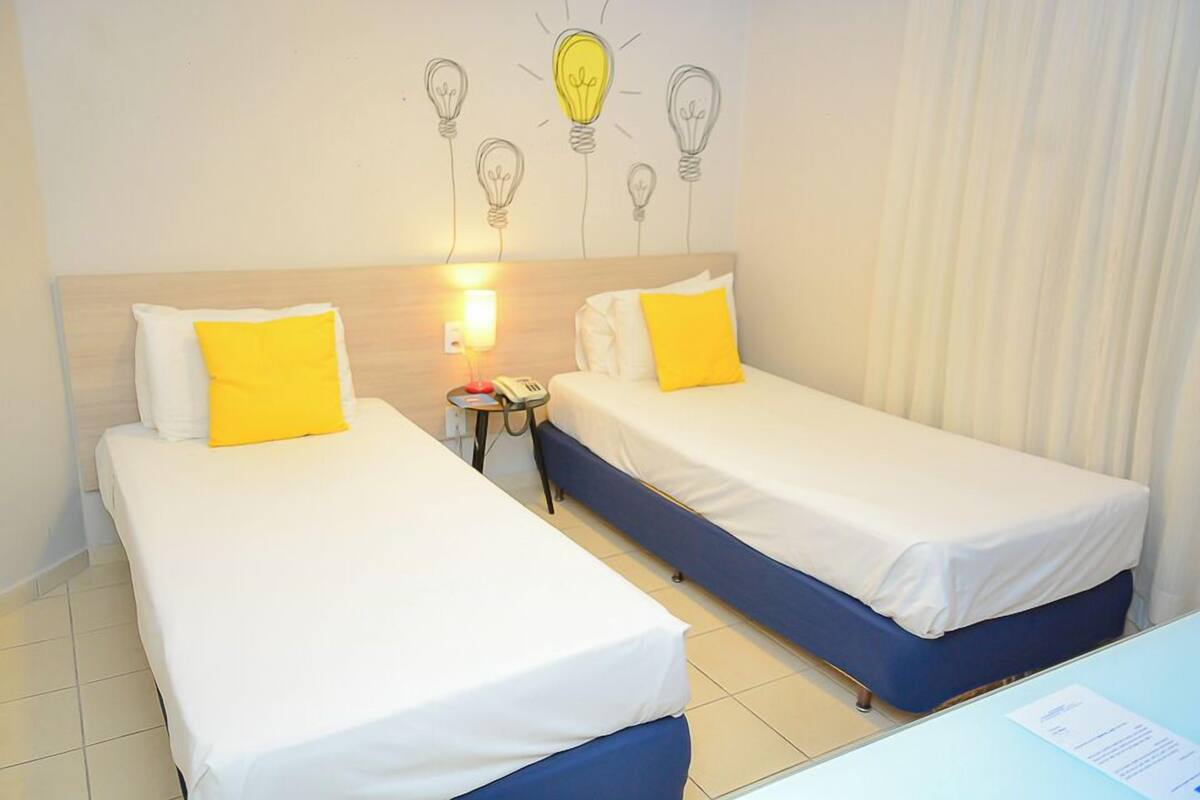 Habitación de hotel con dos camas, cada una con una colcha blanca y un cojín amarillo. Hay una mesita de noche con una lámpara y un teléfono. En la pared, un dibujo de una bombilla.