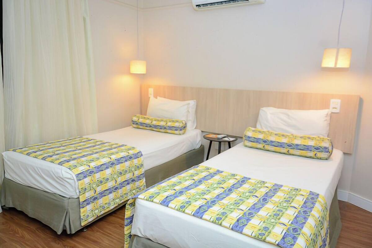 Habitación de hotel con dos camas individuales, cada una con una colcha estampada y almohadas blancas. Hay una mesita de noche pequeña con una lámpara y un equipo de aire acondicionado en la pared.