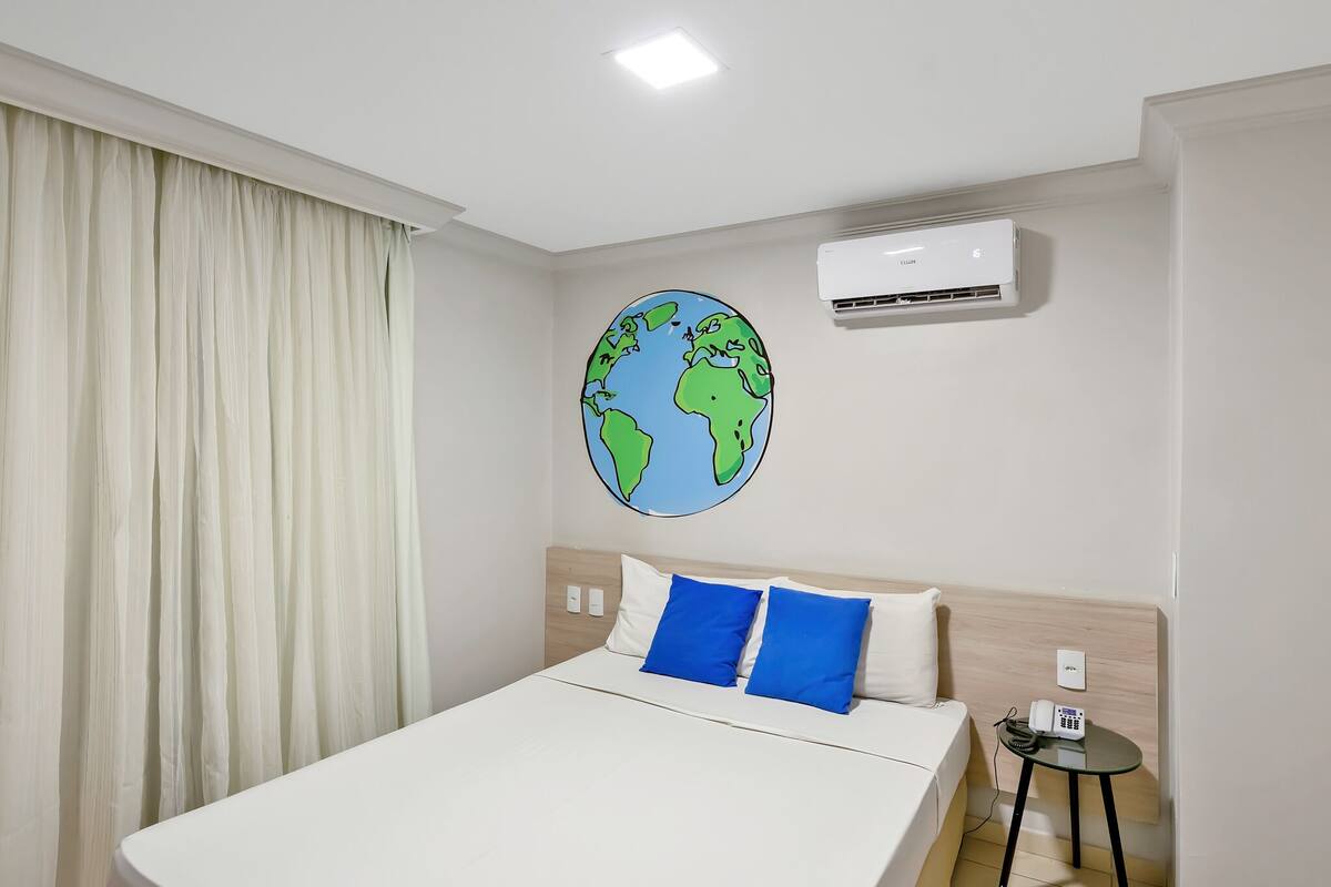 Una habitación de hotel con cama, un adorno redondo en la pared representando la Tierra, un equipo de aire acondicionado, una mesita de noche con teléfono y una cortina.