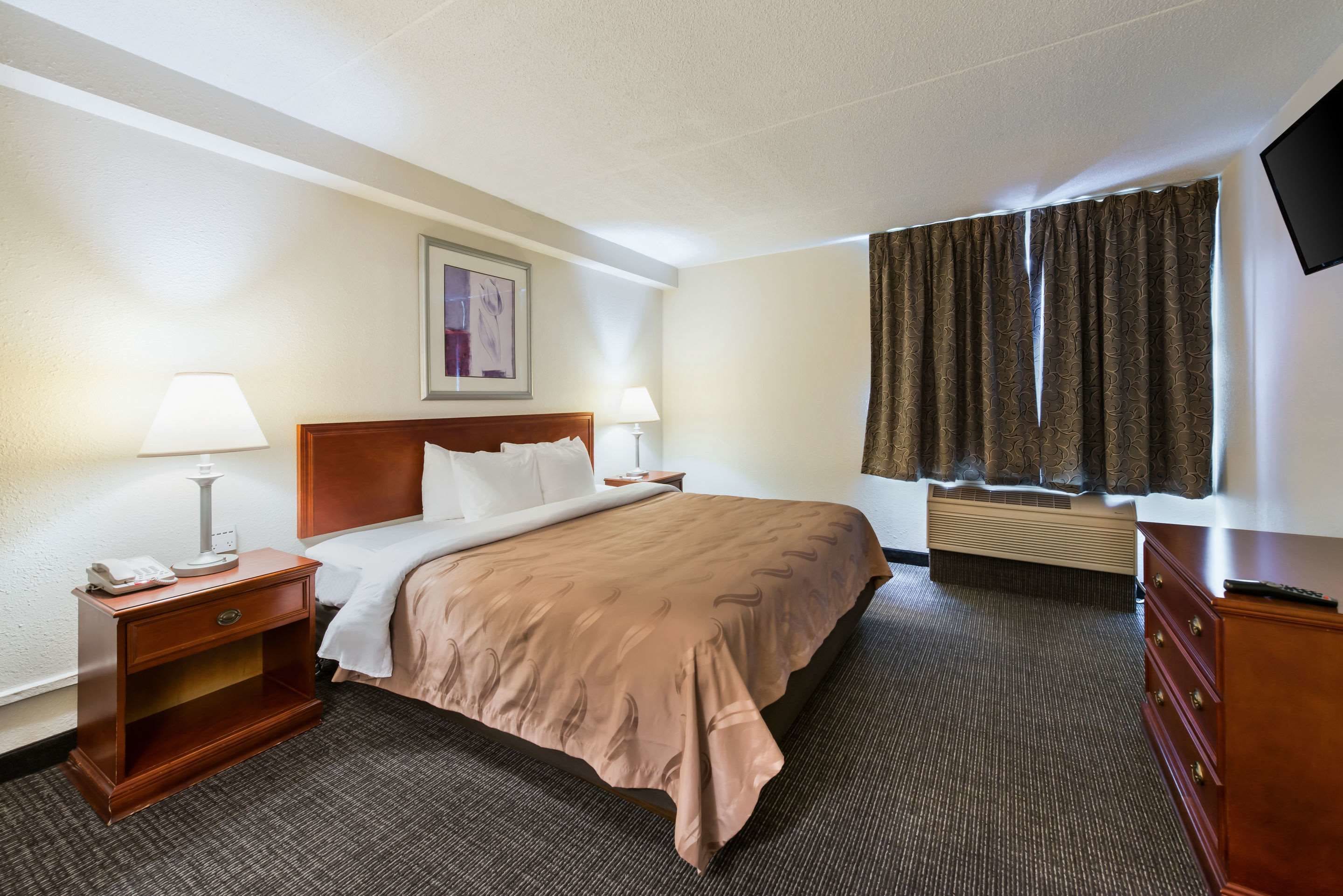 A hotel room with a bed, bedside lamps, a nightstand, a dresser, a flat-screen TV, and a window with curtains.