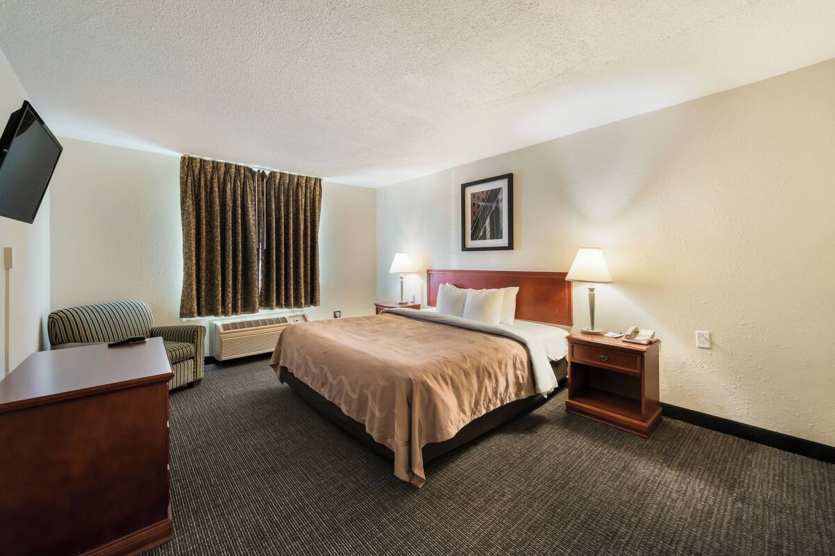 A hotel room with a bed, a desk, a chair, a TV, a window with curtains, and a lamp.