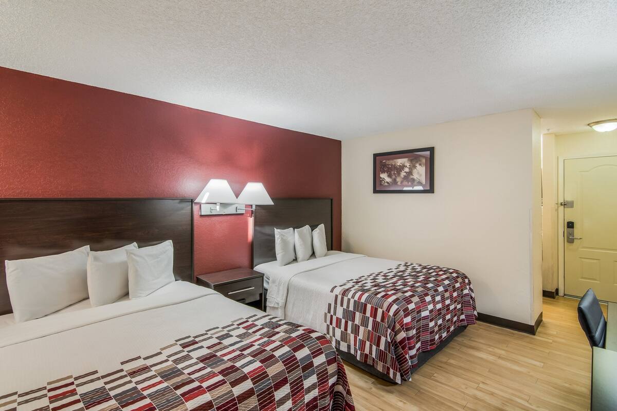 Habitación de hotel con dos camas, una mesita de noche con lámparas, un cuadro enmarcado en la pared y una puerta a la derecha.