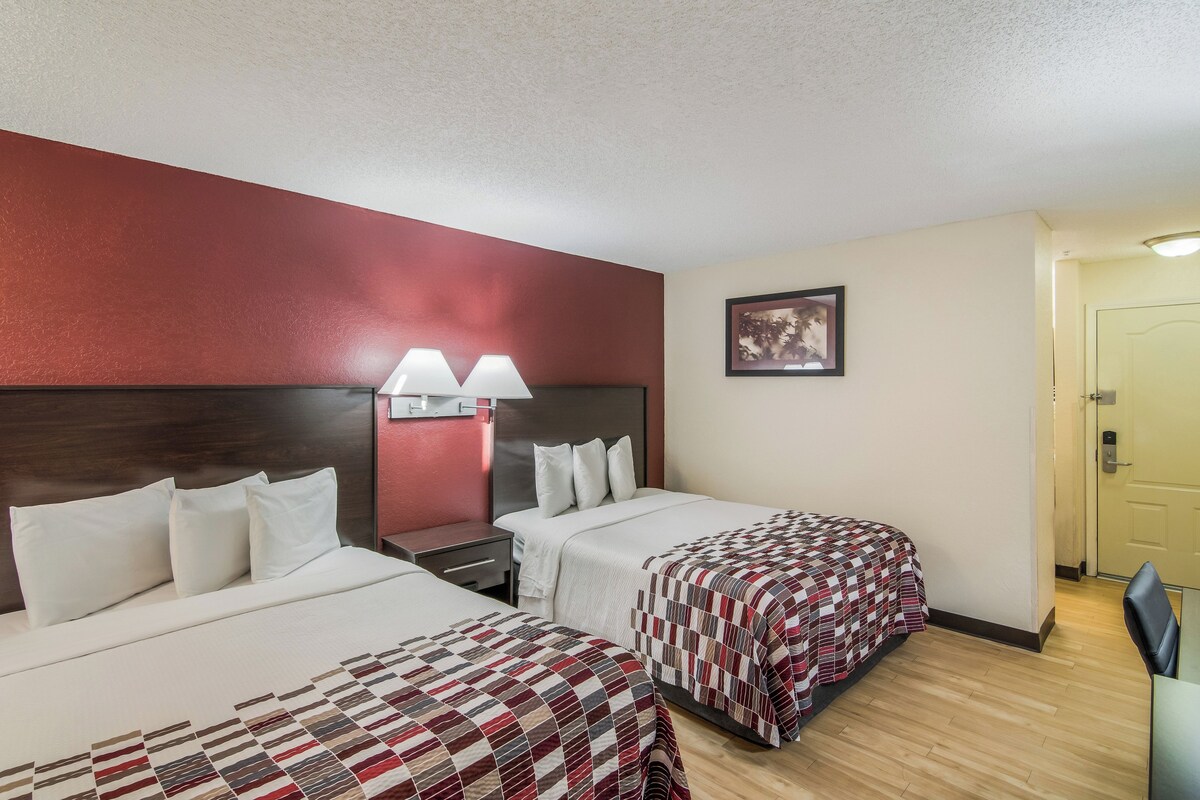 A hotel room with two beds, a nightstand with lamps, a framed picture on the wall, and a door to the right.