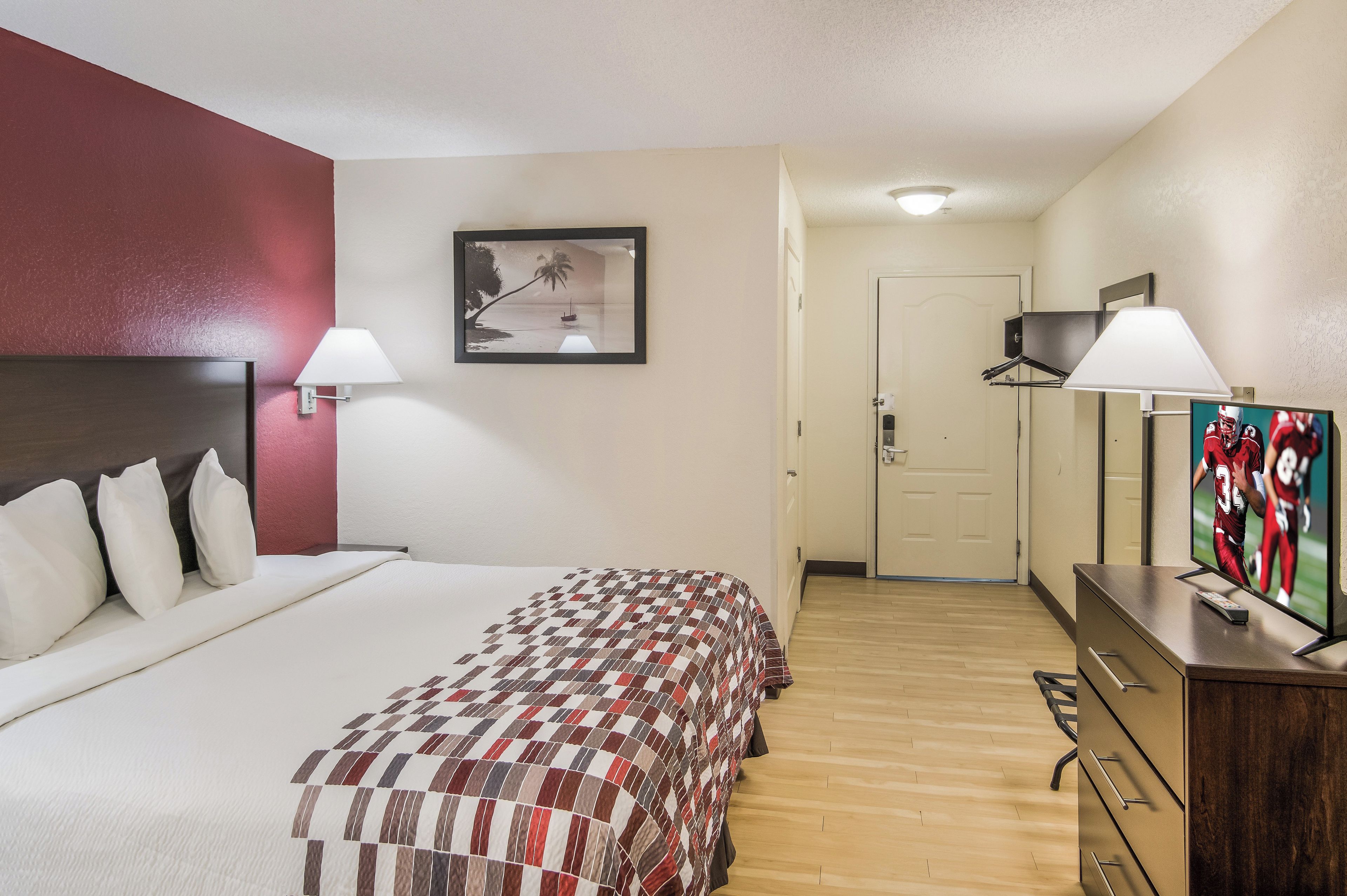 A hotel room with a bed, a desk with a TV, a door, and a wall-mounted picture.