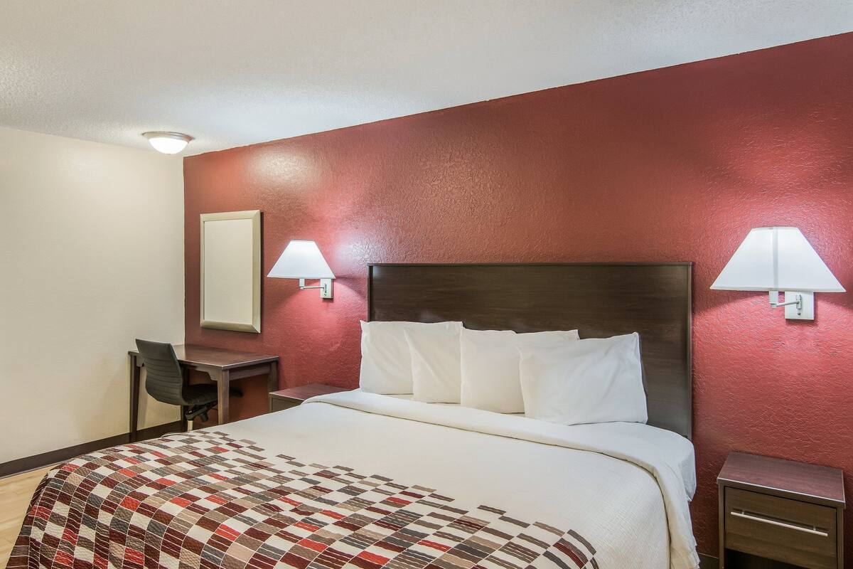 Una habitación de hotel con cama, escritorio, silla y dos lámparas fijadas a la pared.