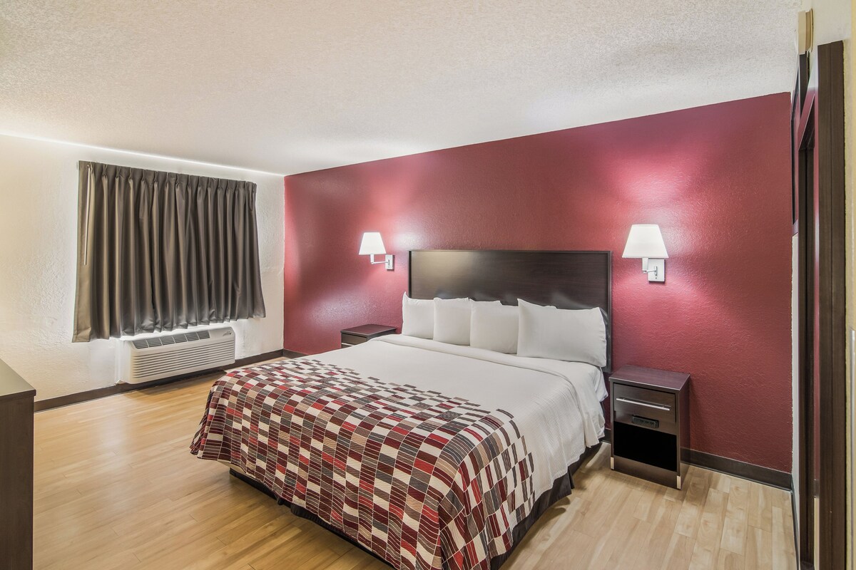 A hotel room with a bed, two bedside tables, a window with curtains, and a wall-mounted lamp.