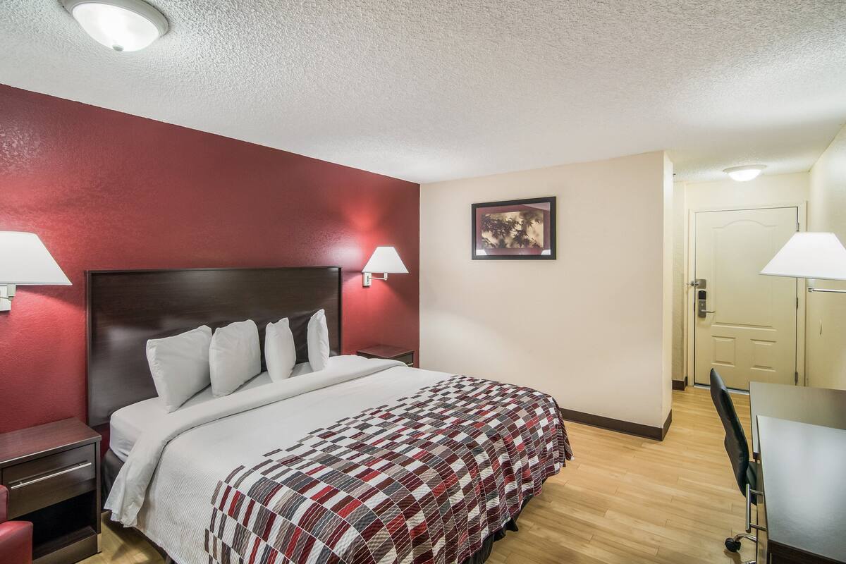 Habitación de hotel con una cama grande, dos lámparas de noche, cabecero de madera, pared de acento roja, un cuadro enmarcado y un escritorio con silla.