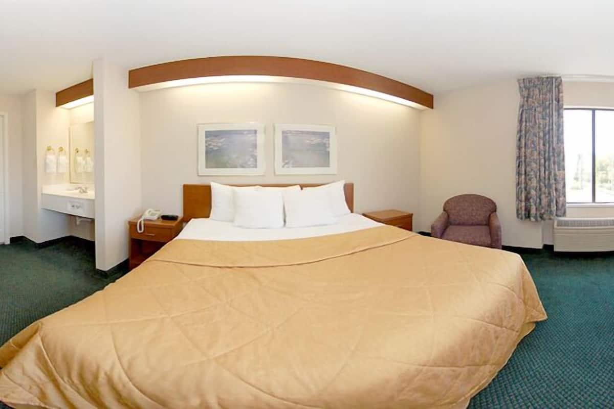 Una habitación de hotel con una cama grande, una mesita de noche con lámpara, una silla, una ventana con cortinas y un teléfono montado en la pared.