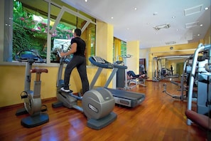 A gym with treadmills, ellipticals, and weight machines, large windows with a view of greenery, and a wooden floor. - Hotel Caparena (Taormina)