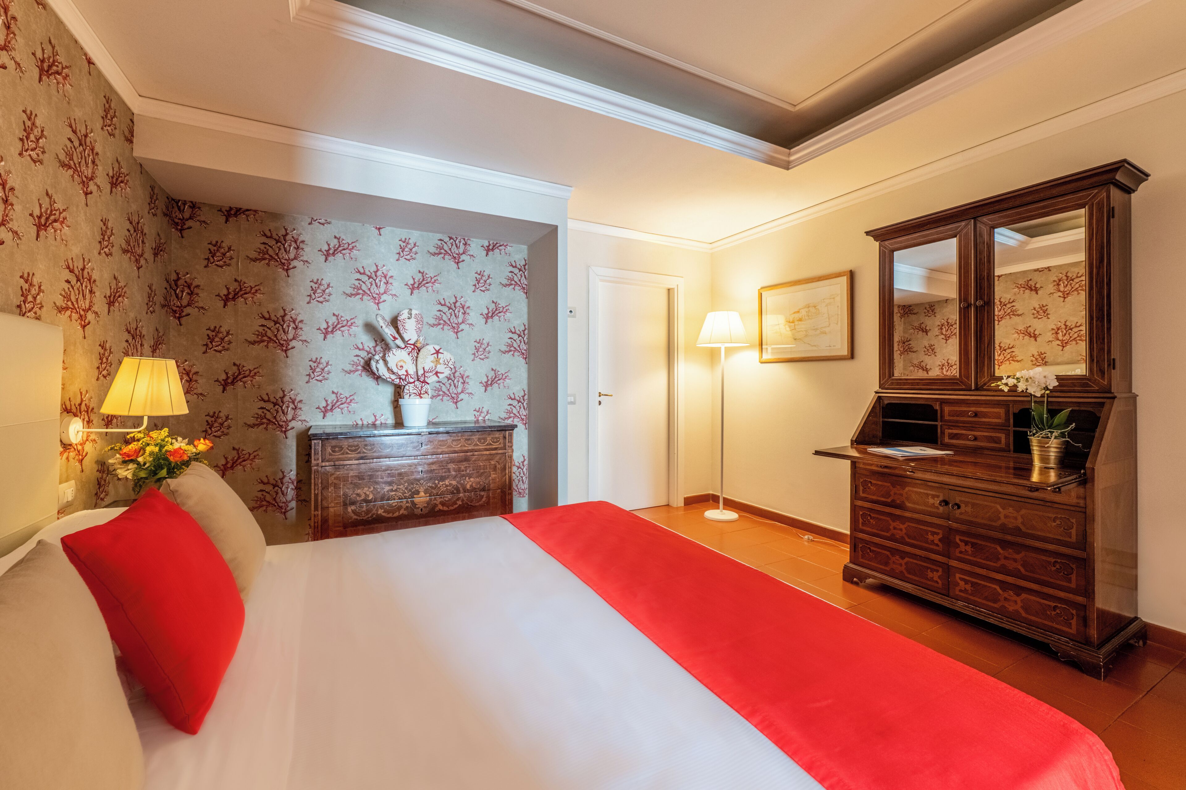 A hotel room with a bed, dresser, lamp, and floral wallpaper.