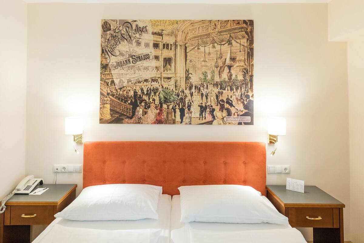 A hotel room with a large poster of a historical opera house, a tufted orange headboard, two bedside tables with lamps, and a telephone on the left table.
