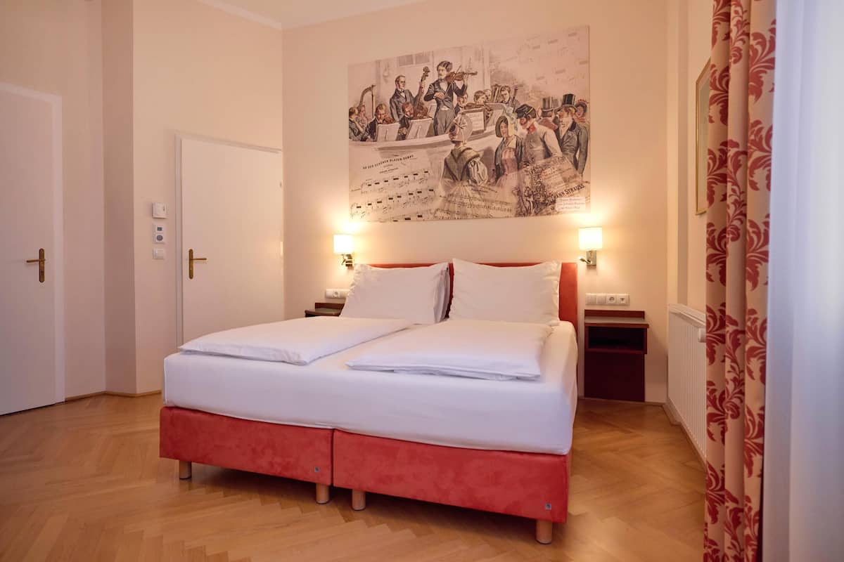 A double bed with white linens, a red headboard, two bedside tables with lamps, a wall-mounted poster, and a wooden floor.