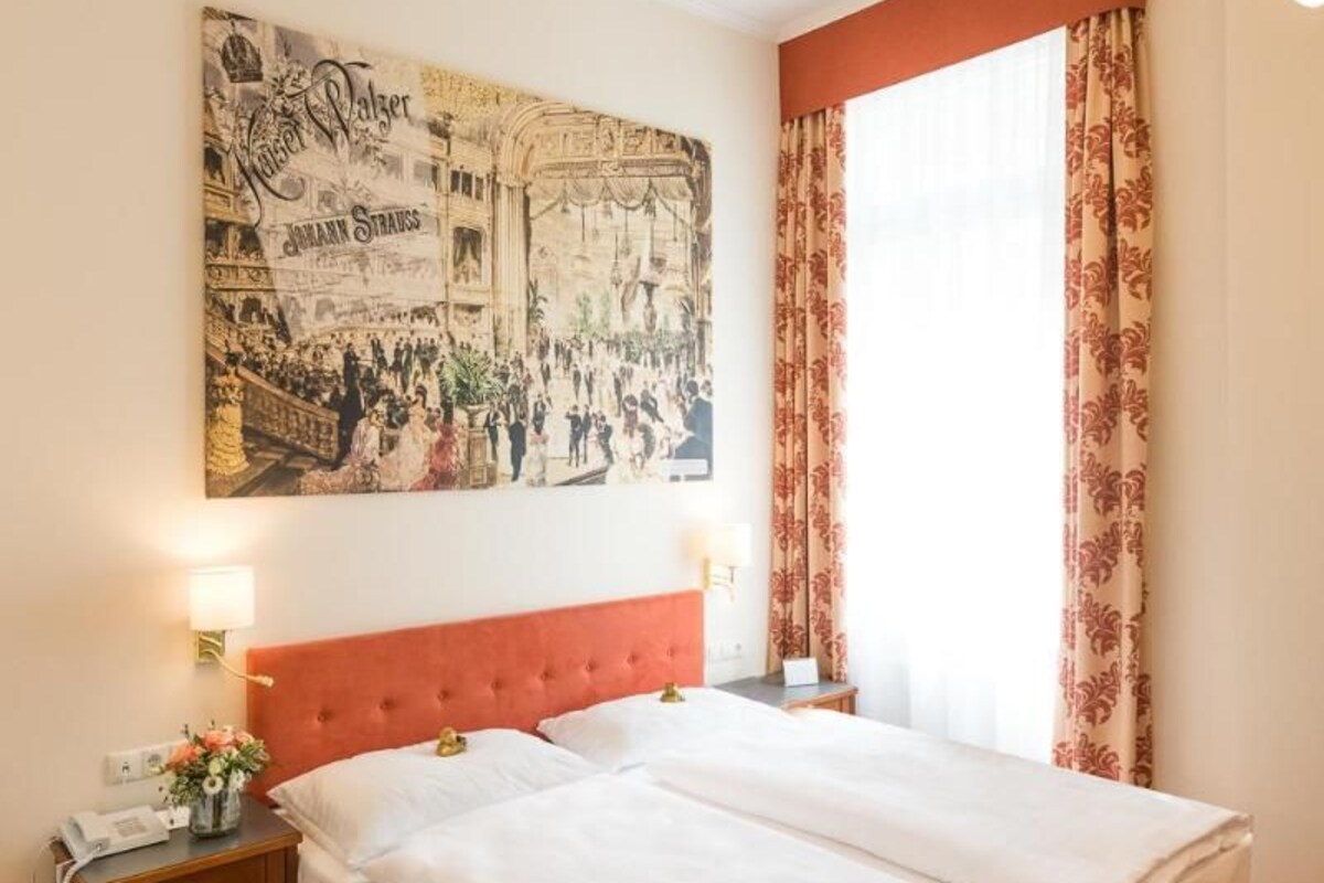 A double bed with a red headboard, a nightstand with a lamp, a wall-mounted artwork, and a window with curtains.