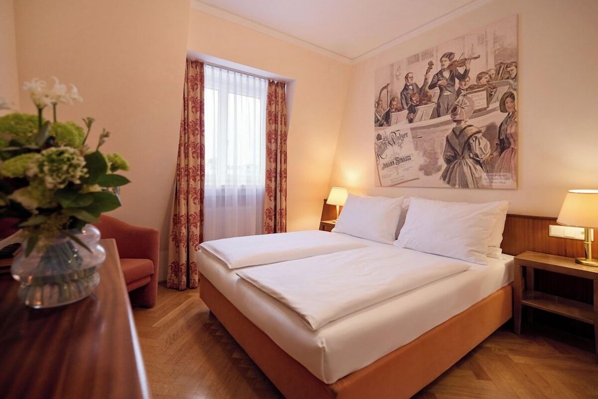 A hotel room with a double bed, a wooden nightstand, a desk with a vase of flowers, a red chair, and a large poster on the wall.