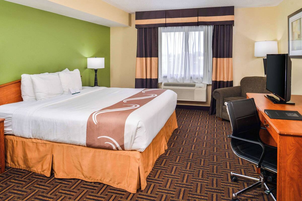 A hotel room with a large bed, a desk with a computer, a chair, a television, and a window with curtains.