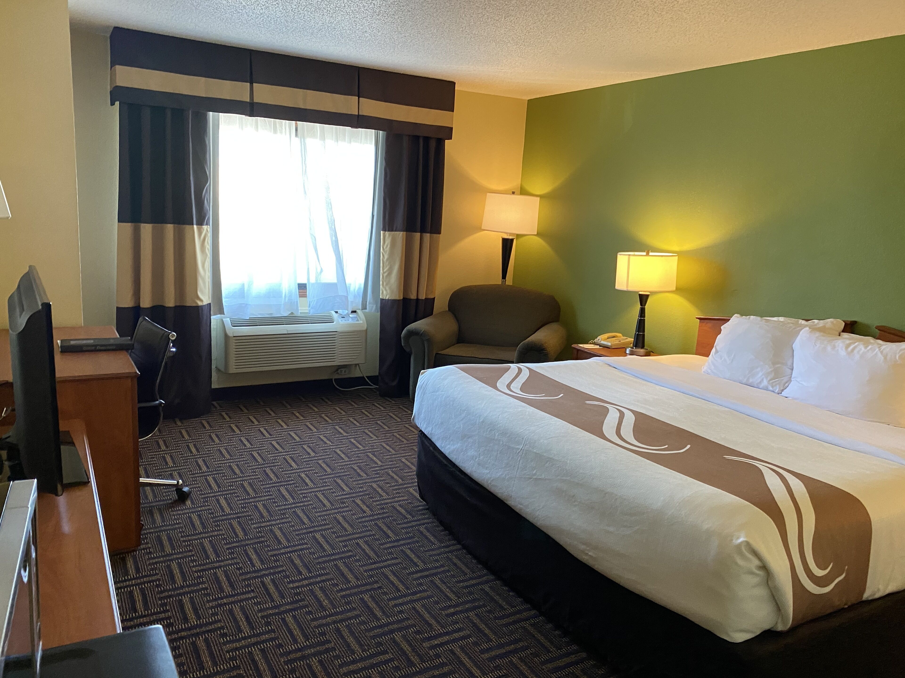 A hotel room with a bed, desk, chair, air conditioning unit, and two lamps.