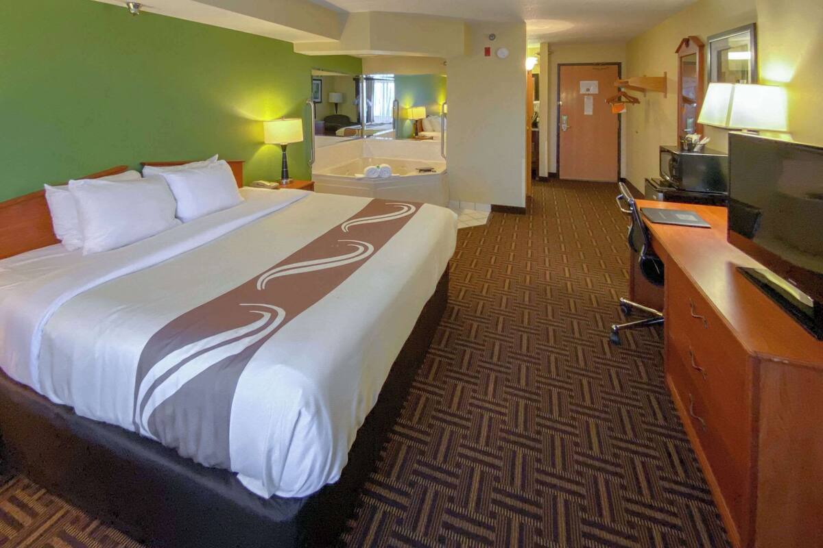 A hotel room with a large bed, a desk with a television, a bathroom with a bathtub, and a carpeted floor.
