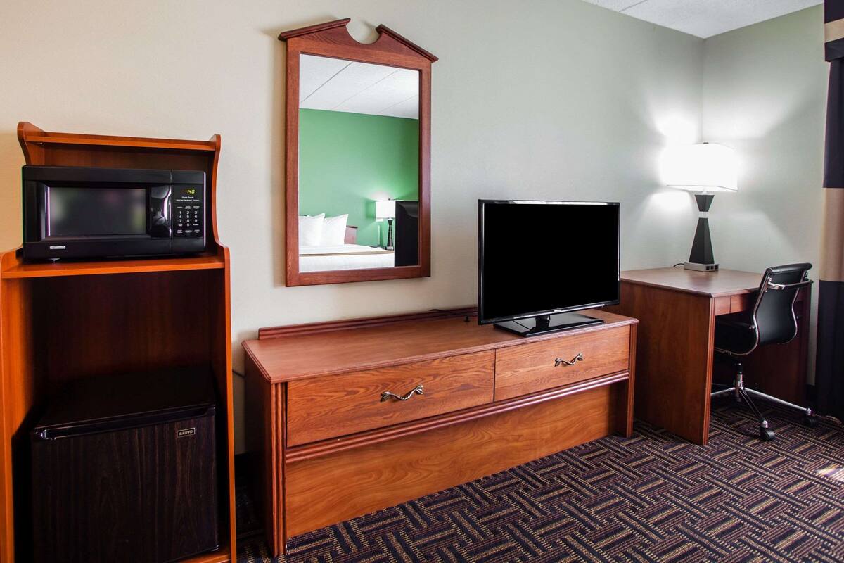 A hotel room with a wooden dresser, a microwave, a flat-screen TV, a desk with a lamp, and a chair.
