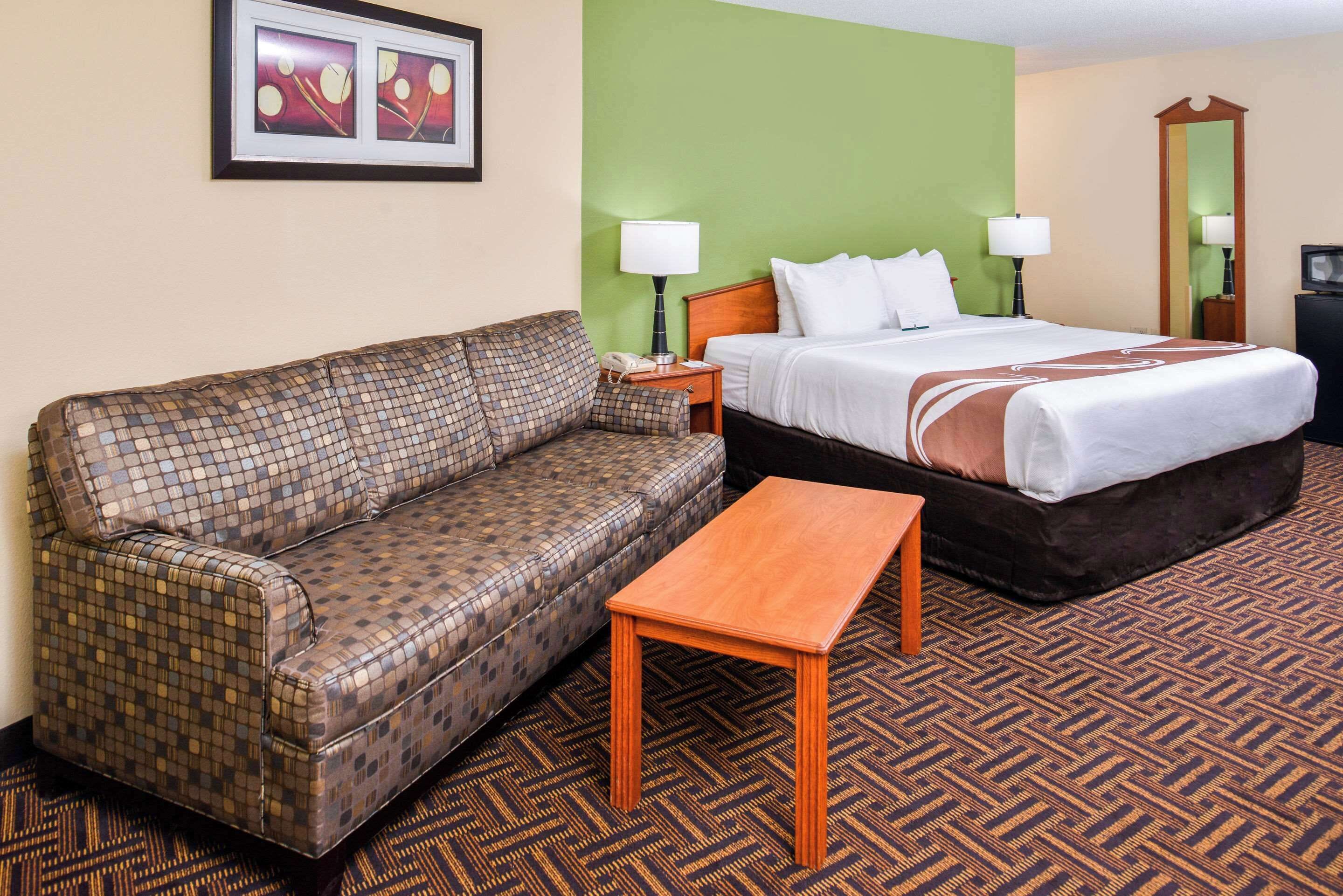 A hotel room with a checkered sofa, a bed with white bedding, a wooden nightstand, and a patterned carpet.