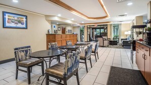 A hotel lobby with a reception desk, seating area with tables and chairs, and a view of the outside.