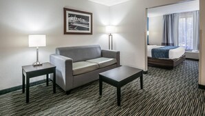 A hotel room with a grey sofa, a small table, a lamp, a framed picture on the wall, and a bed with a blue blanket. - Comfort Inn Gainesville - University Area (Gainesville)
