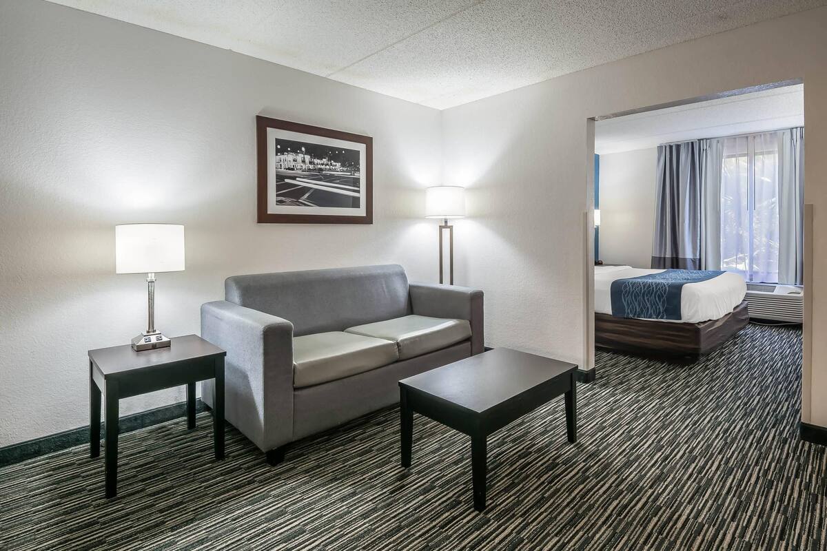 Quarto de hotel com sofá cinzento, uma mesinha, candeeiro, quadro emoldurado na parede e cama com manta azul.