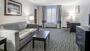 A hotel room with a grey sofa, a small table, a desk, a chair, and a large window with curtains. - Comfort Inn Gainesville - University Area (Gainesville)