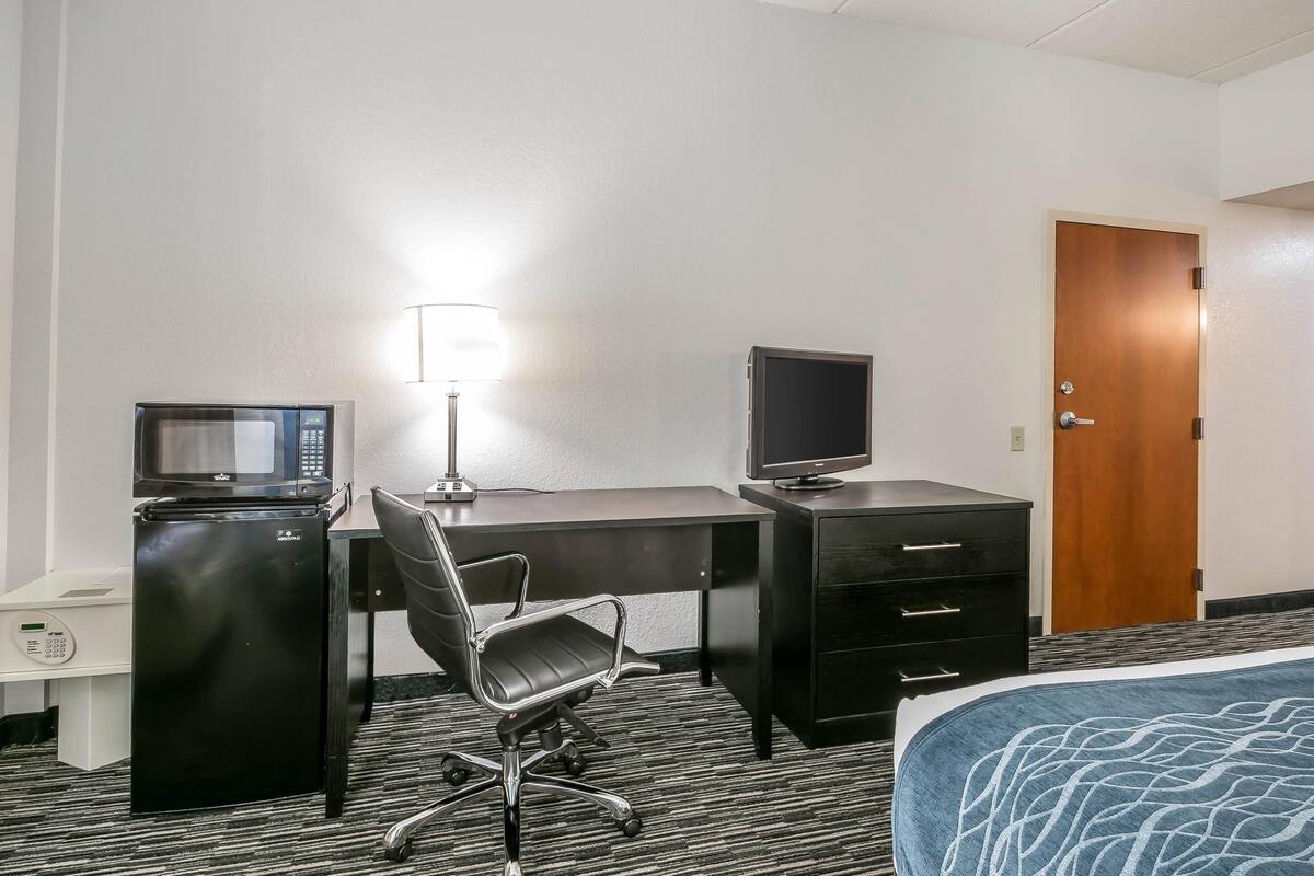 Quarto de hotel com escrivaninha, computador, televisão, micro-ondas, frigorífico, cadeira, cama e porta.